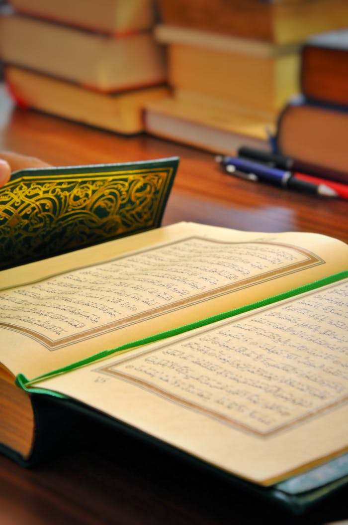 Close-up of an open Quran on a wooden table with books and a pen, symbolizing study and reflection.